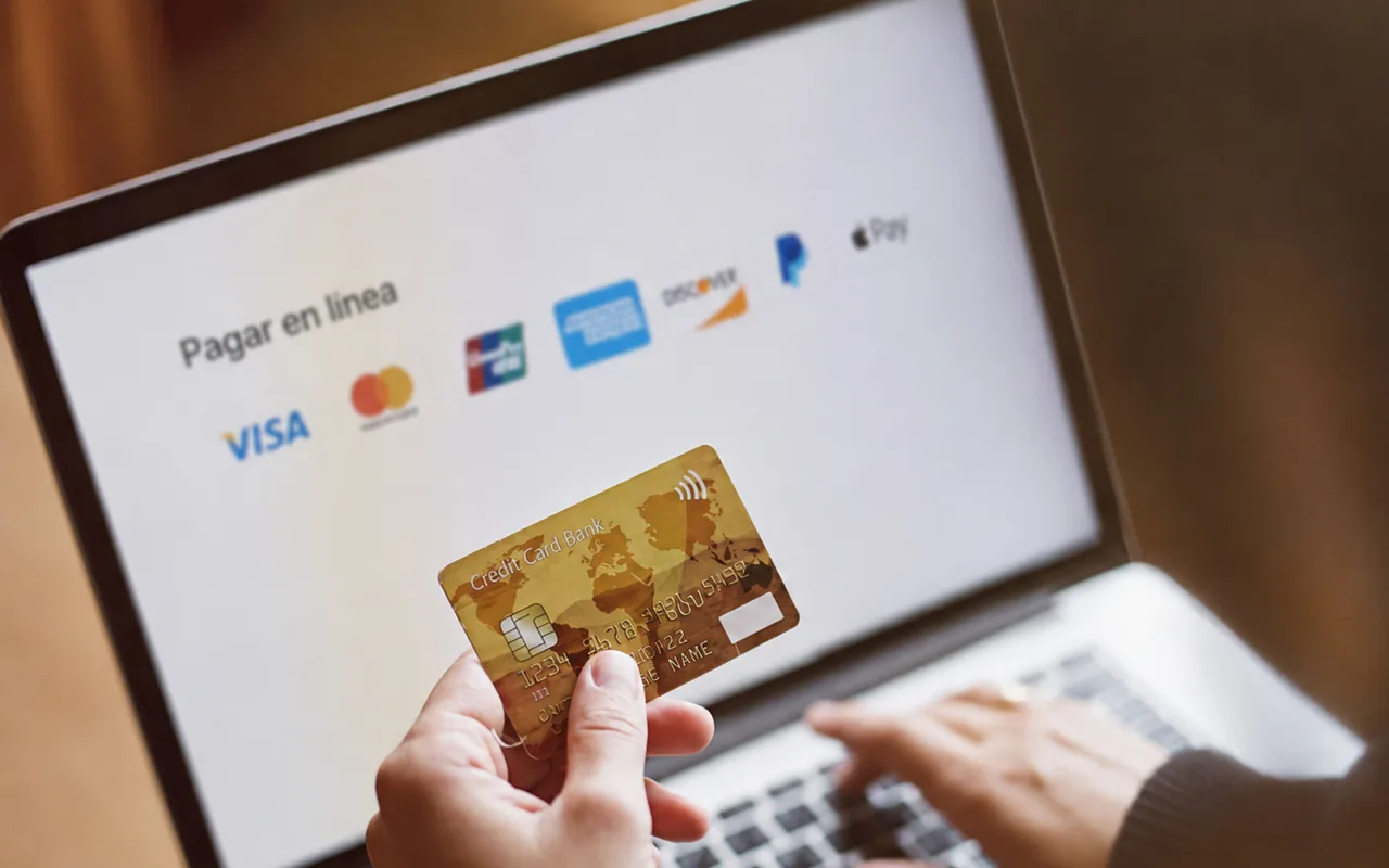 Cliente sostiene su tarjeta de crédito frente a la pantalla de pago en línea de una página web, con los logotipos de las principales tarjetas y medios de pago.